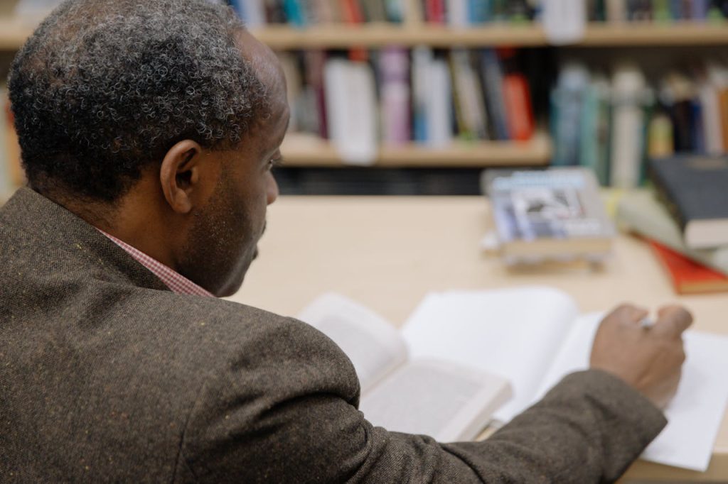 Man in Brown Suit Jacket Reading and Writing Inside a Library